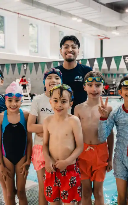 A swim coach stands with six smiling children in swimwear and goggles at an Asphalt Green indoor pool, posing together near the edge of the water.