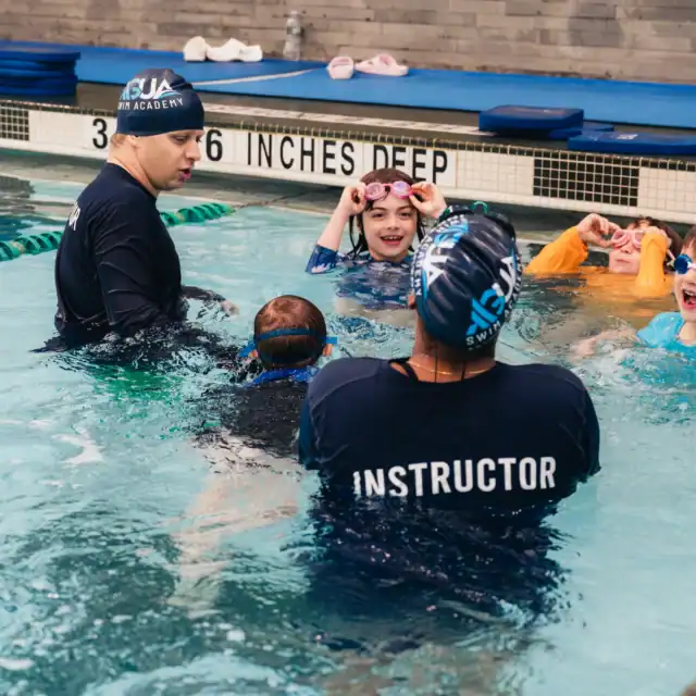 Two swimming instructors and four children wearing goggles are standing together in a pool during a sports and swim programs lesson.