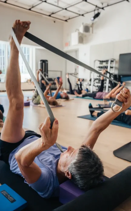 People lying on yoga mats in a bright studio at Asphalt Green use straps to stretch their legs during a group fitness class. Yoga blocks and equipment are visible around them.