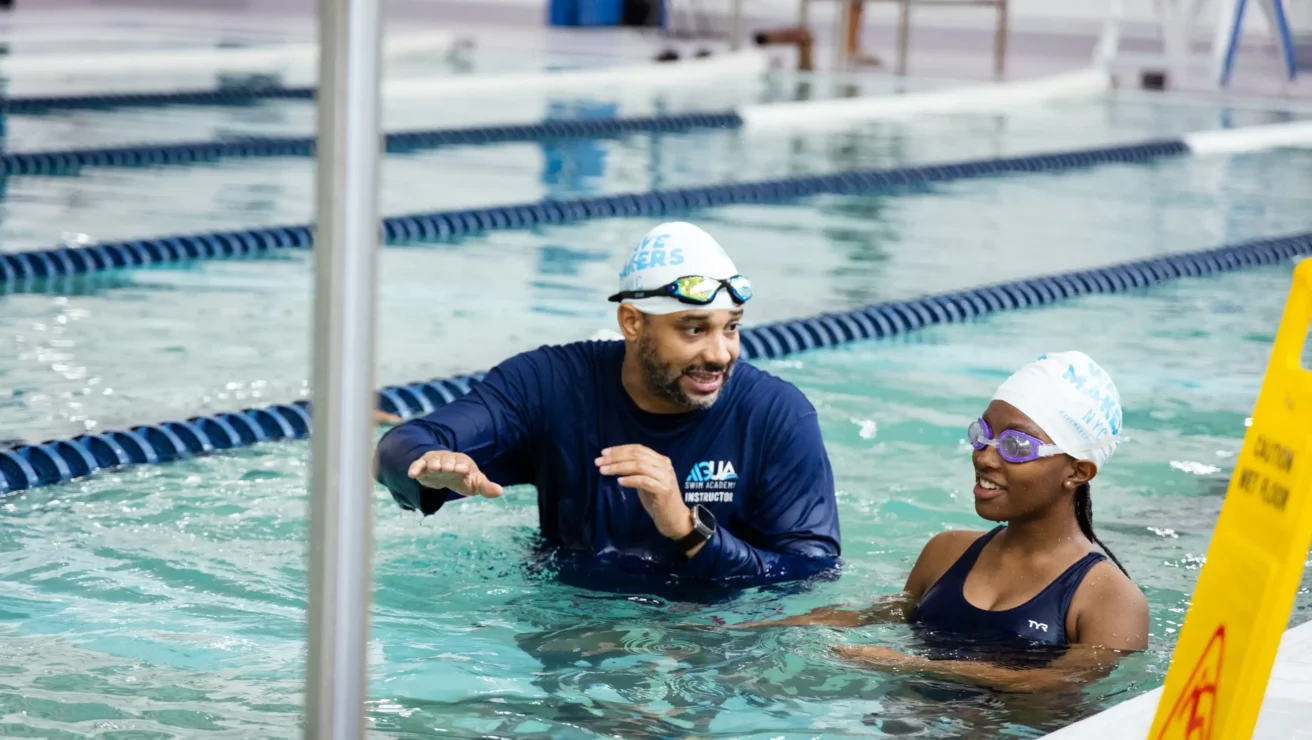 A swim instructor in a blue shirt talks to a young swimmer in goggles and a swim cap about water safety in an indoor pool near lane dividers.