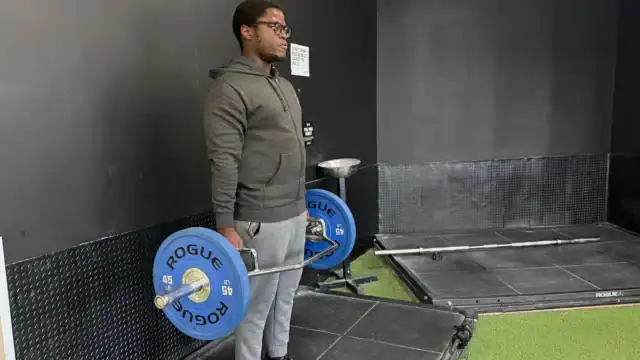 Person wearing glasses and workout clothes stands in a gym holding a barbell with blue Rogue weight plates, facing forward, with black walls and gym equipment in the background.