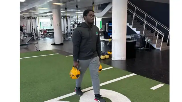 A man in athletic wear walks on indoor turf holding two yellow kettlebells, with gym equipment and stairs visible in the background.