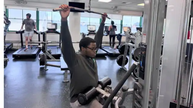 A person uses a lat pulldown machine in a gym, with others running on treadmills in the background.