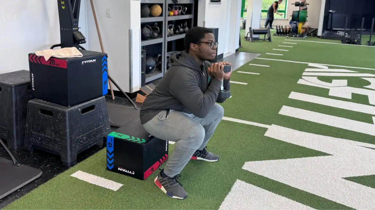 Person in athletic wear performs a box squat exercise with a dumbbell on artificial turf in a gym setting, showcasing longevity training techniques aimed at promoting strength and overall well-being.