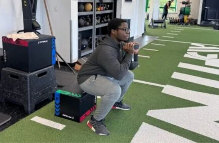 Person in athletic wear performs a box squat exercise with a dumbbell on artificial turf in a gym setting, showcasing longevity training techniques aimed at promoting strength and overall well-being.