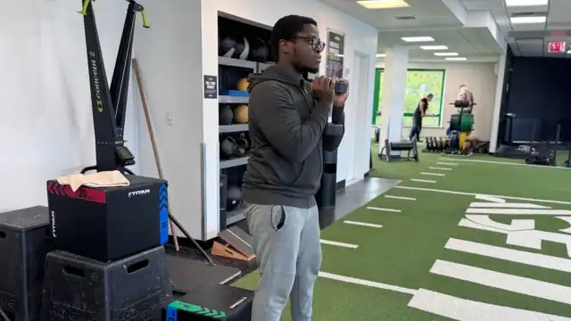 A person in athletic wear holds a weight close to their chest, practicing longevity training in a gym with turf flooring, equipment, and shelves with medicine balls in the background.