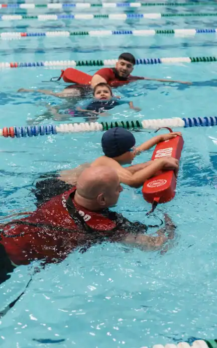 Two adults wearing red lifeguard shirts assist two children with flotation devices during a swimming lesson at Asphalt Green’s indoor pool.