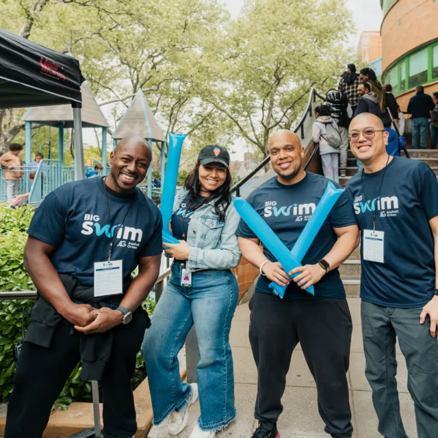 Four people stand outdoors smiling at a community event at Asphalt Green, wearing “Big Swim” shirts and holding blue foam sticks; playground and building in the background.