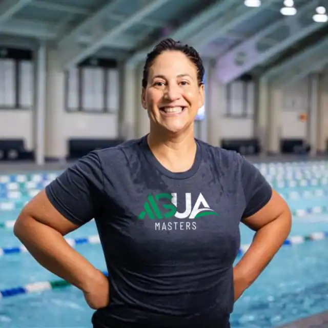 Person in a dark t-shirt with “AQUA Masters” logo stands poolside in an indoor swimming facility, smiling at the camera, highlighting the positive ag impact of community-driven aquatic programs.