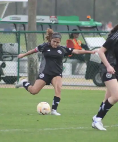 Soccer player in black uniform prepares to kick a ball during a game on a grassy field, with other players and equipment visible in the background.