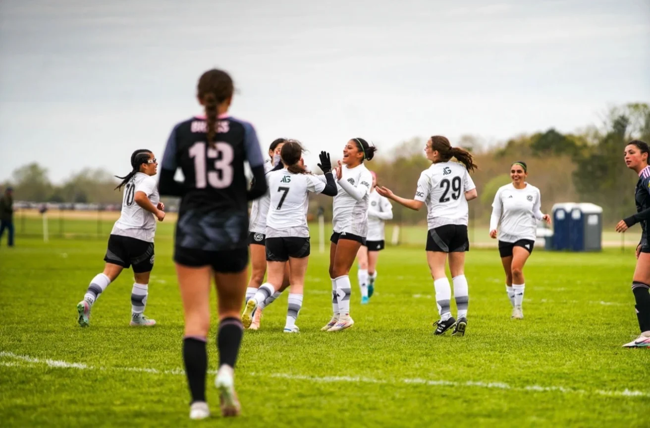 A group of soccer players in white jerseys celebrate on a field as a player in a black jersey with number 13 stands in the foreground.