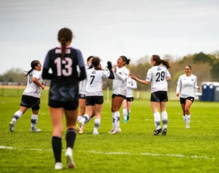 A group of soccer players in white jerseys celebrate on a field as a player in a black jersey with number 13 stands in the foreground.