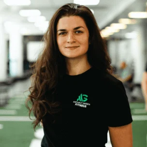 Woman with long brown hair wearing a black Asphalt Green Fitness t-shirt stands in a brightly lit gym with a neutral expression.
