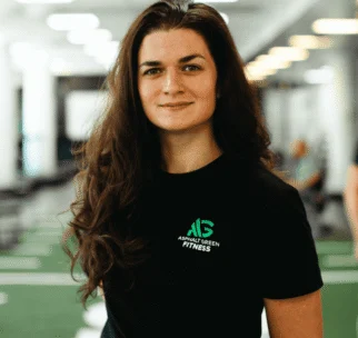 Woman with long brown hair wearing a black Asphalt Green Fitness t-shirt stands in a brightly lit gym with a neutral expression.