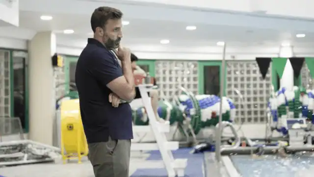 A man stands poolside indoors, arms crossed, looking thoughtful. Pool equipment, a yellow bin, and swimming lanes are visible in the background.