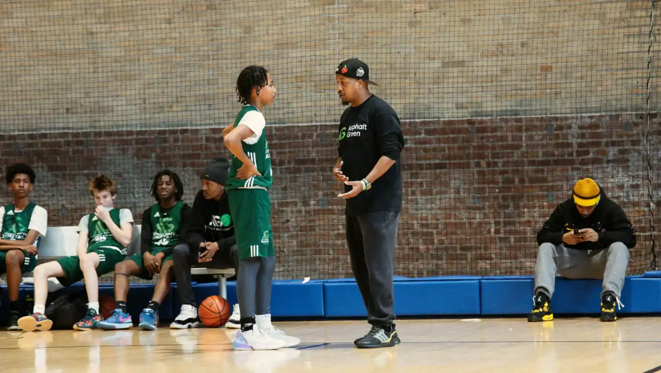 A basketball coach talks to a young player on the court while other team members sit on the bench; one person on the end is looking at a phone, perhaps checking out tips about water safety for their upcoming team outing.