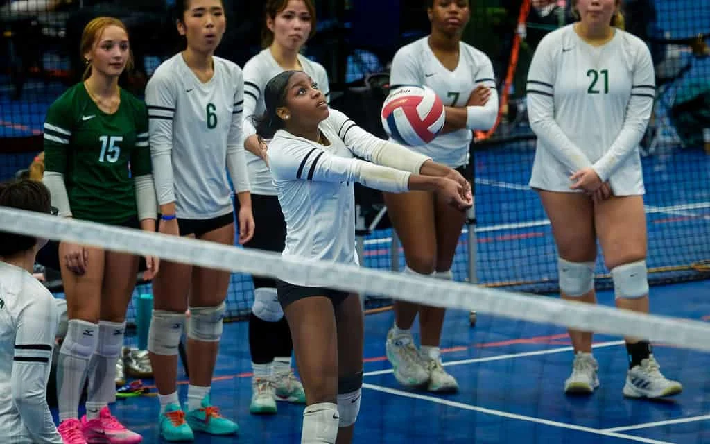 A volleyball player in a white jersey bumps the ball during a game while teammates and opponents watch from behind the net.