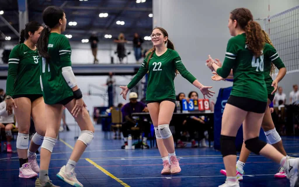 Five volleyball players in green jerseys celebrate together on an indoor court, with a scoreboard in the background showing scores of 13 and 19.