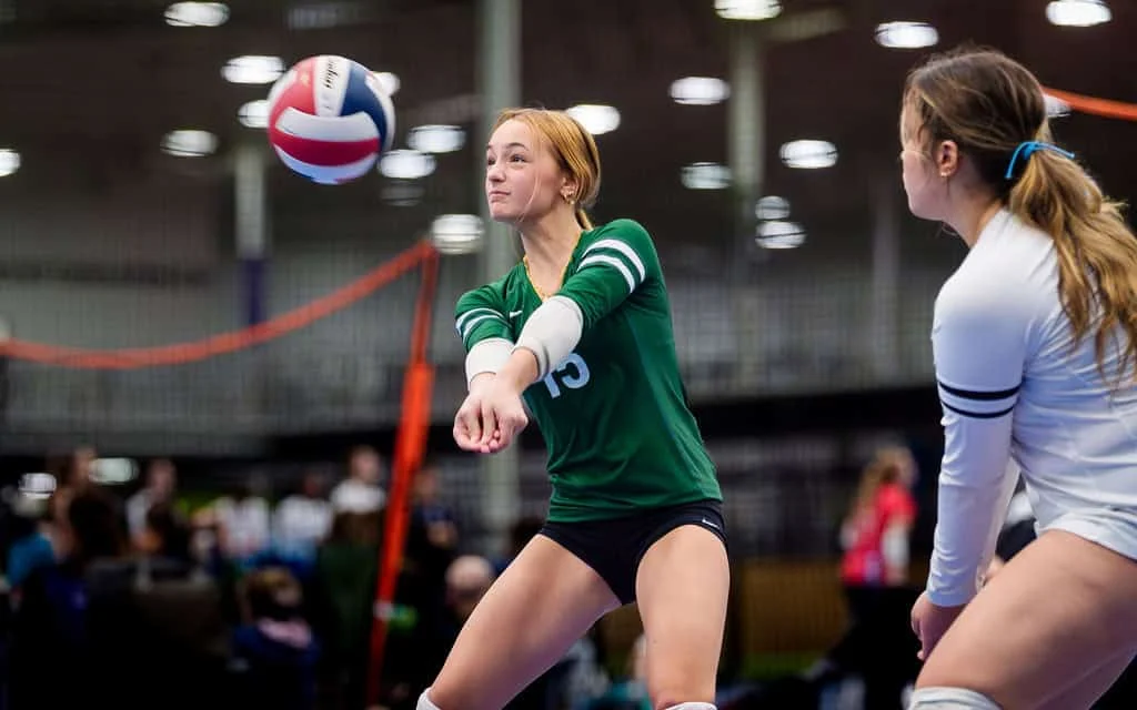 A volleyball player in a green jersey bumps the ball, while another player in white watches during an indoor game.