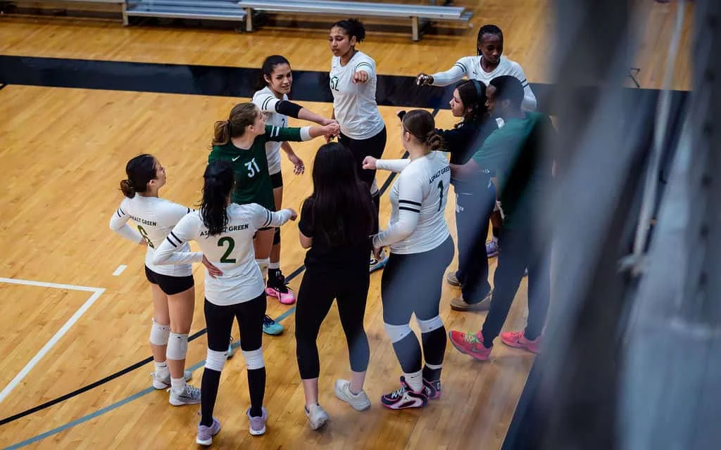 A women’s volleyball team and coaches stand in a circle on a gym floor, joining their hands together, preparing for a game or practice.