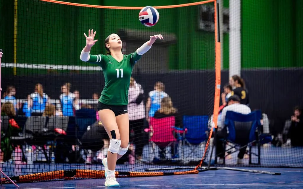 A volleyball player in a green jersey and knee pads serves the ball during an indoor match; spectators sit in the background.