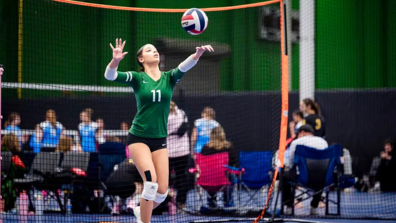 A volleyball player in a green jersey and knee pads serves the ball during an indoor match; spectators sit in the background.