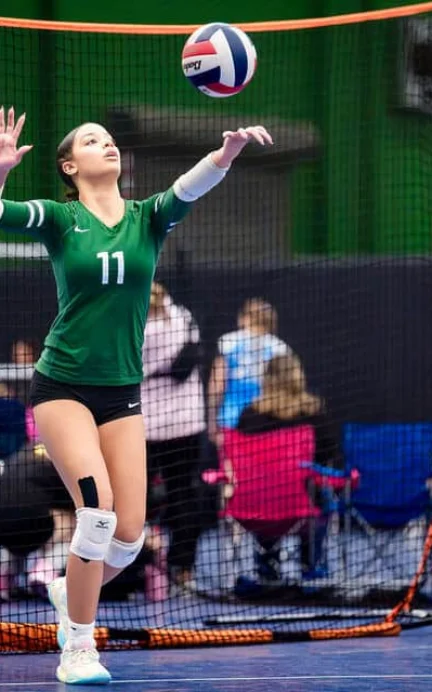 A volleyball player in a green jersey and knee pads serves the ball during an indoor match; spectators sit in the background.