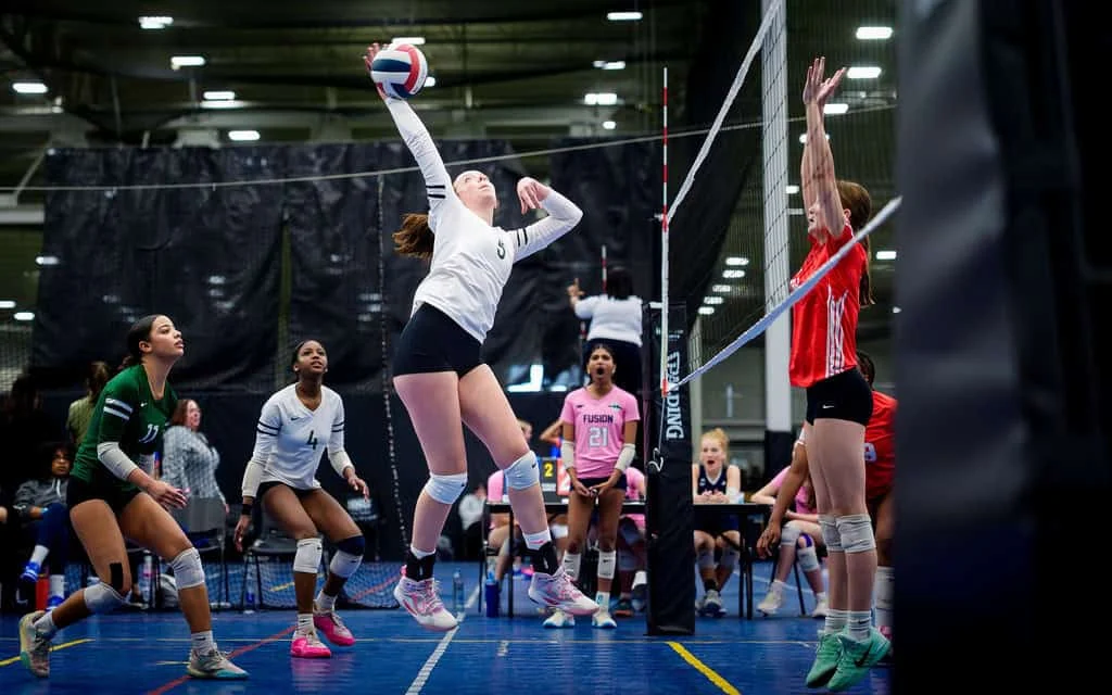 A volleyball player in a white jersey jumps to spike the ball while players from both teams prepare to react during an indoor match.