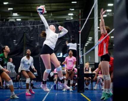 A volleyball player in a white jersey jumps to spike the ball while players from both teams prepare to react during an indoor match.