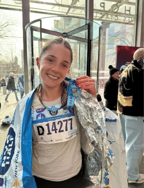 A smiling runner wearing a race bib numbered 14277 and a foil emergency blanket draped over her shoulders, holding a finisher medal.