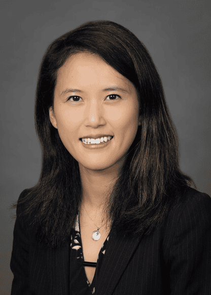A professional headshot of a woman smiling at the camera, wearing a dark pinstripe blazer.