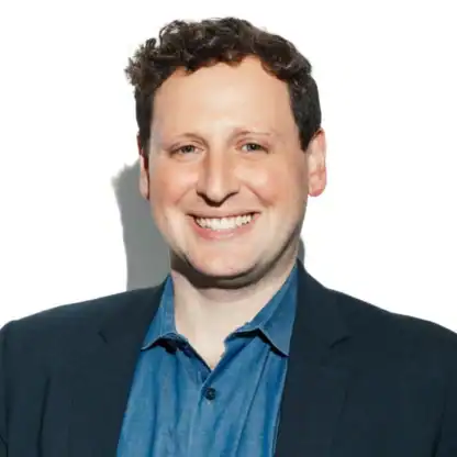 A man in a blue button-down shirt and a dark blazer smiling at the camera.