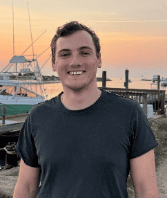 A young man smiling in front of a marina at sunset.