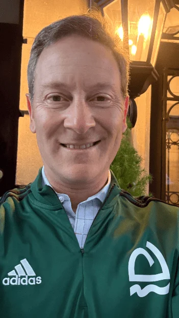 A middle-aged man smiling, wearing a green Adidas zip-up jacket over a collared shirt.
