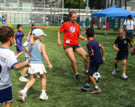 A woman in an England soccer jersey plays soccer with a group of children on an outdoor turf field.