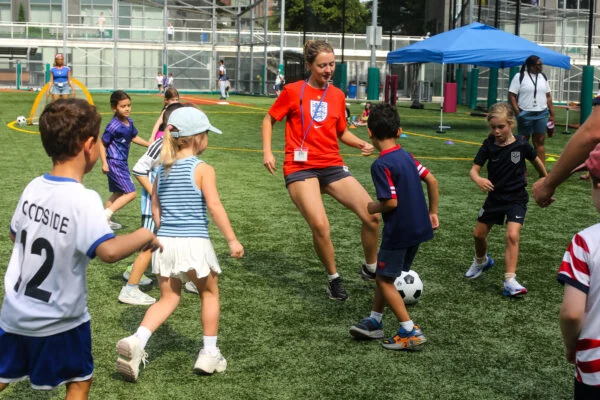 A woman in an England soccer jersey plays soccer with a group of children on an outdoor turf field.
