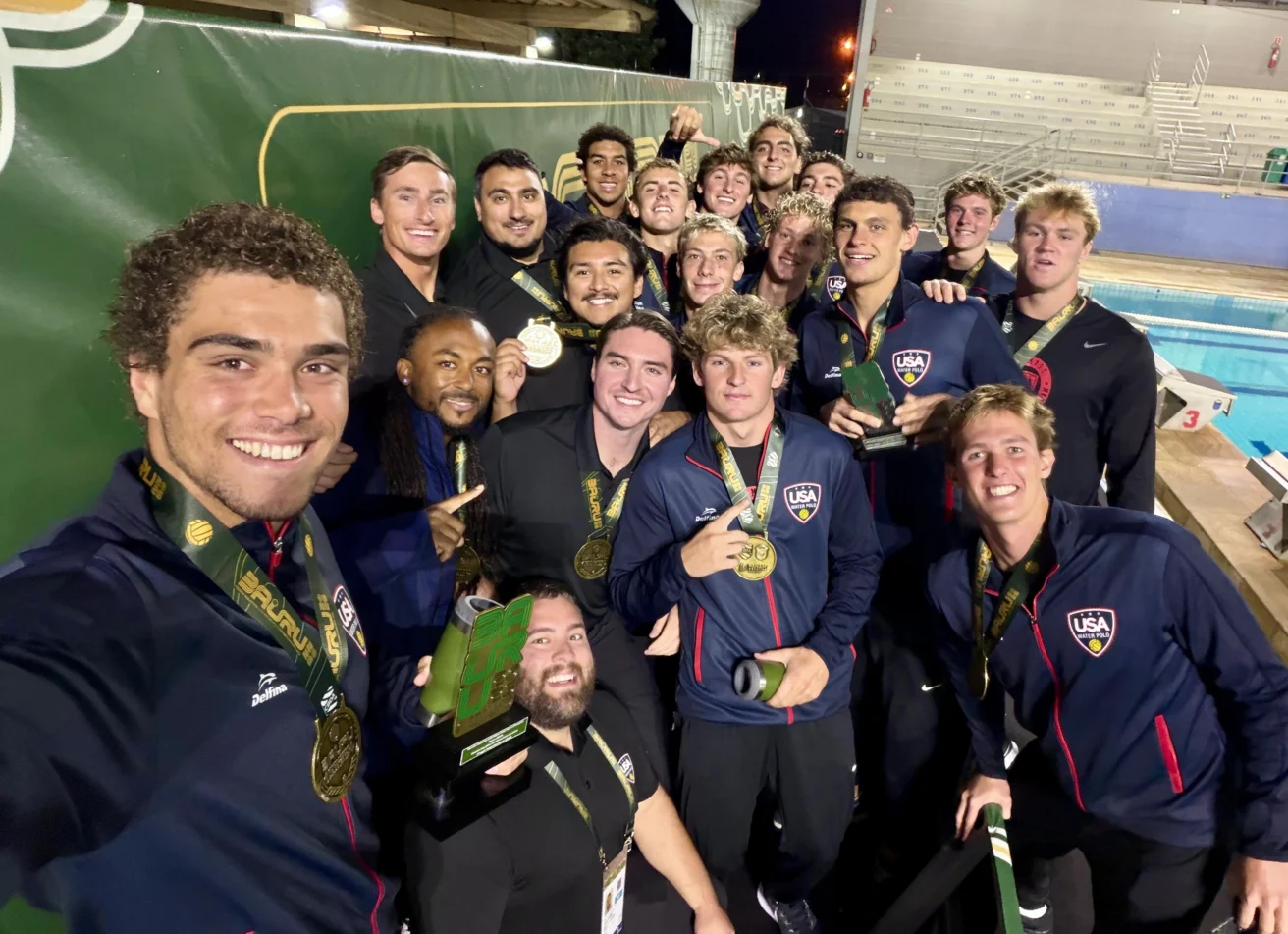 A large group of USA water polo players wearing team jackets and gold medals pose for a selfie by a pool.