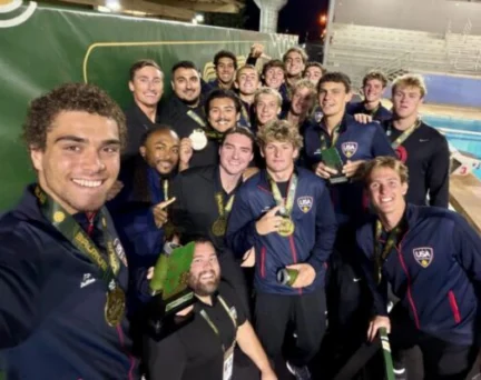 A large group of USA water polo players wearing team jackets and gold medals pose for a selfie by a pool.