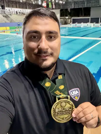 A man wearing a USA Water Polo shirt holds up a gold tournament medal in front of an indoor swimming pool.