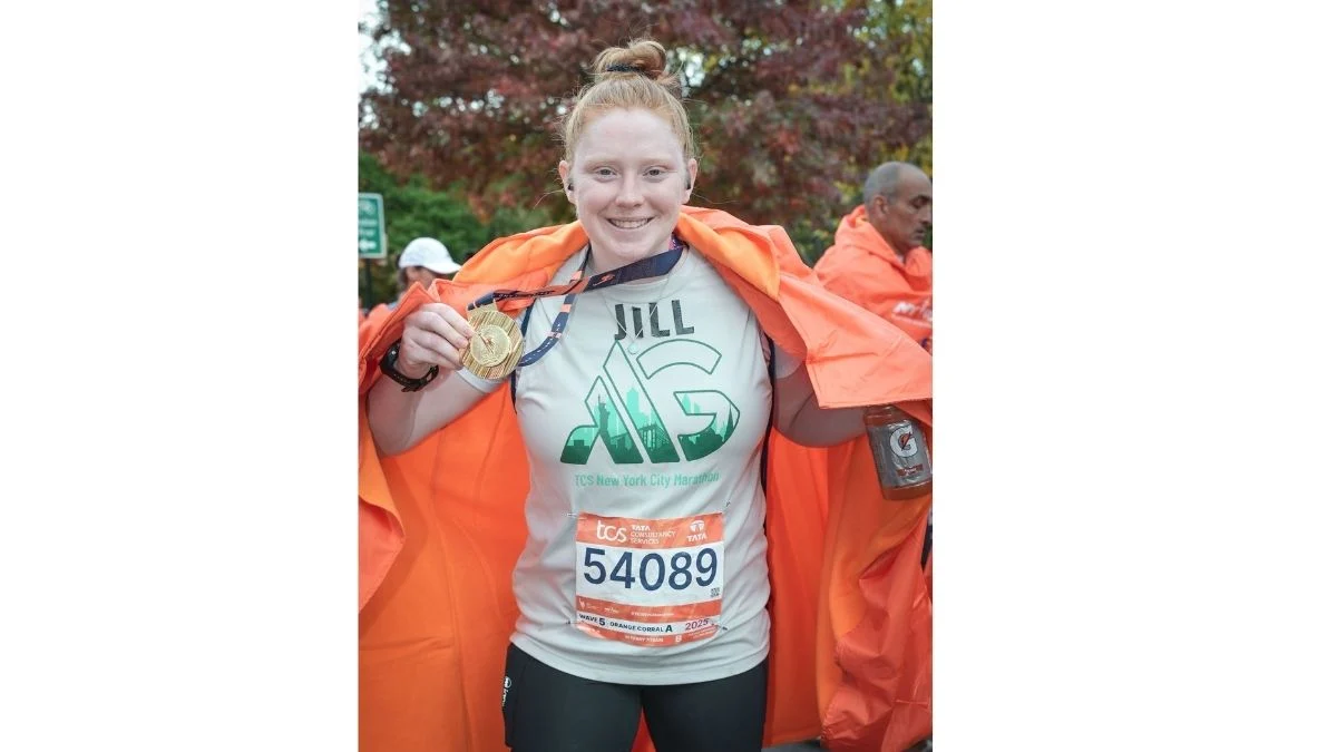 A marathon runner wearing a race bib and a TCS New York City Marathon shirt holds up her finisher medal while draped in an orange cape.