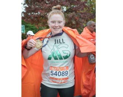 A marathon runner wearing a race bib and a TCS New York City Marathon shirt holds up her finisher medal while draped in an orange cape.