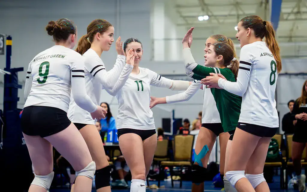 Five female volleyball players on an indoor court celebrate a point, with two players giving high fives.